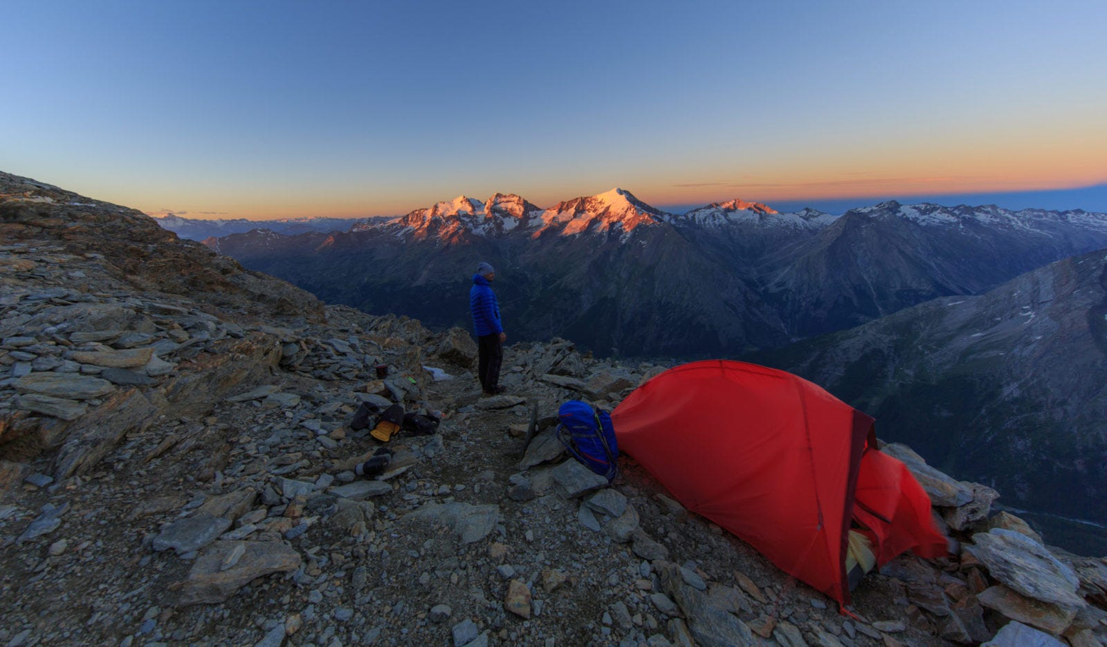 Ein rotes Zelt auf einem felsigen Berg. Im Hintergrund scheint die Sonne auf schneebedeckte Gipfel