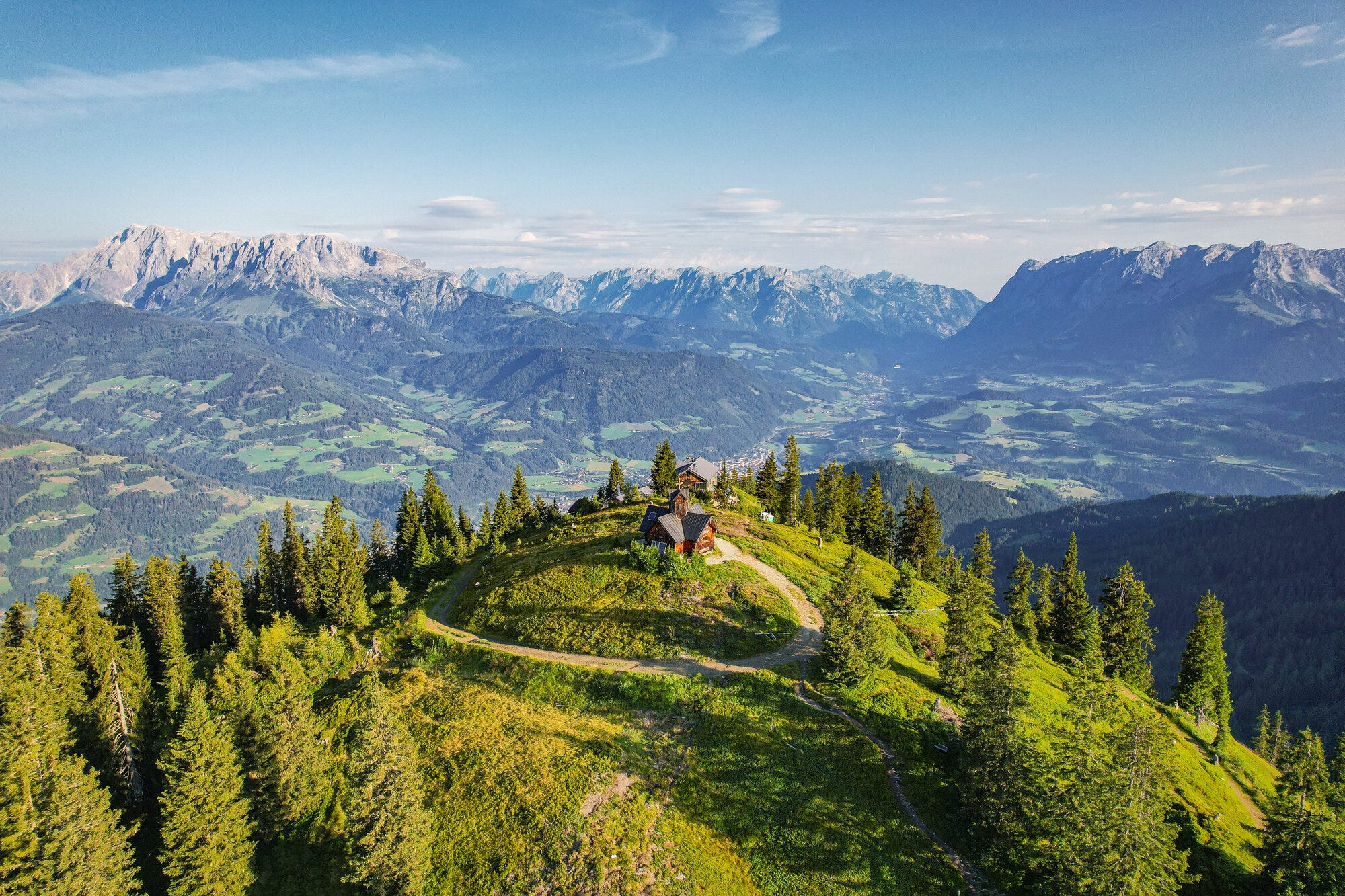 Blick auf einen Hügel mit Hütte und Wanderweg. Dahinter die Bergkulisse von Österreich.