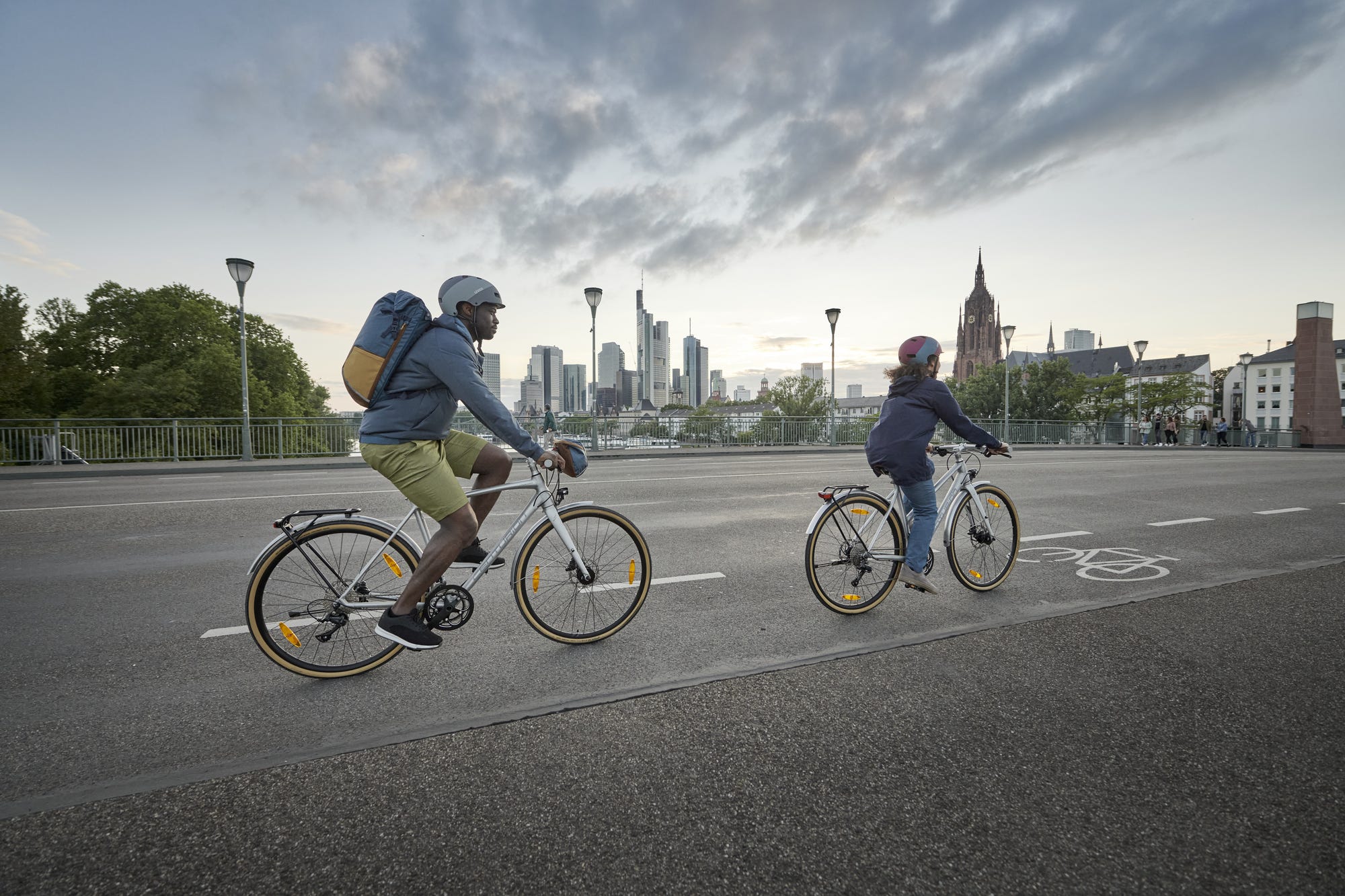 Zwei Radfahrer*innen pendeln im Alltag vor Stadtkulisse
