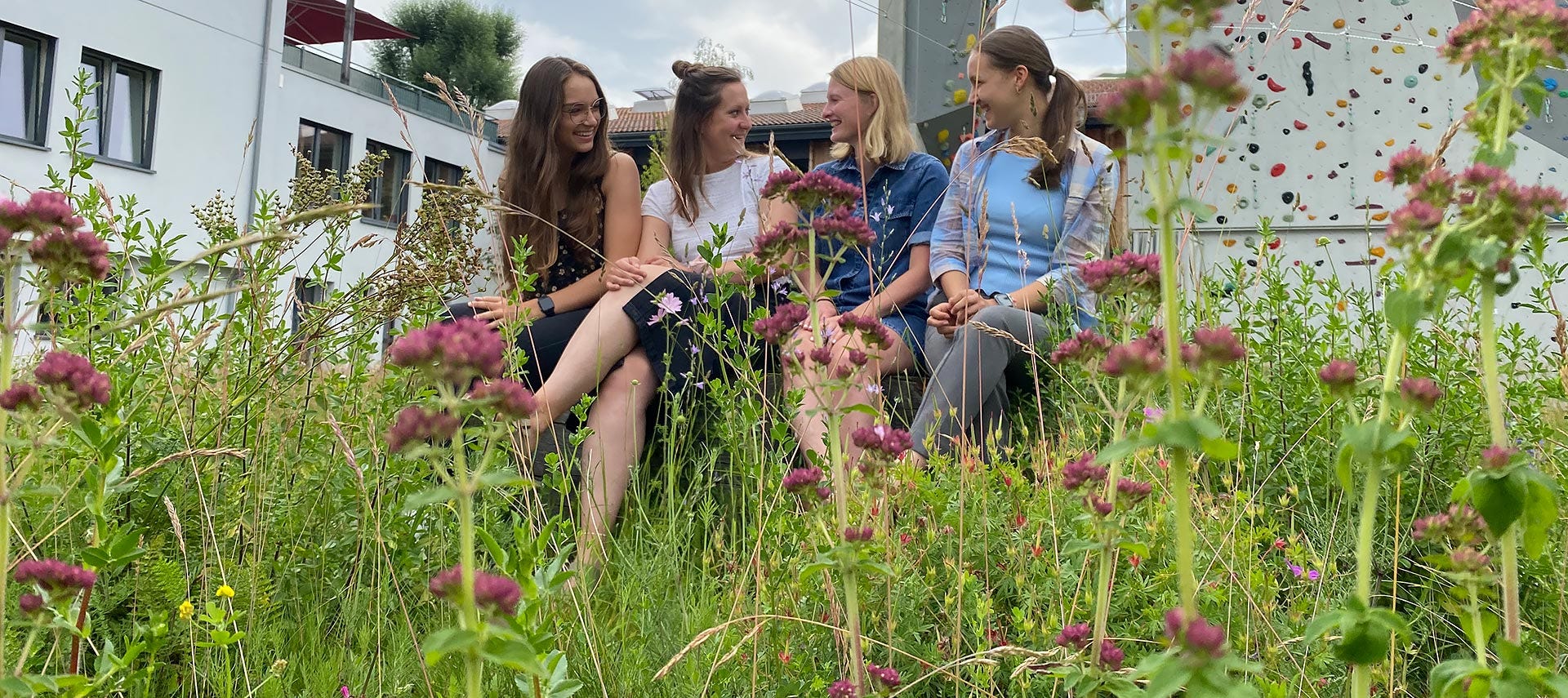 Four VAUDE employees sitting on a bench in the meadow in front of the VAUDE company building