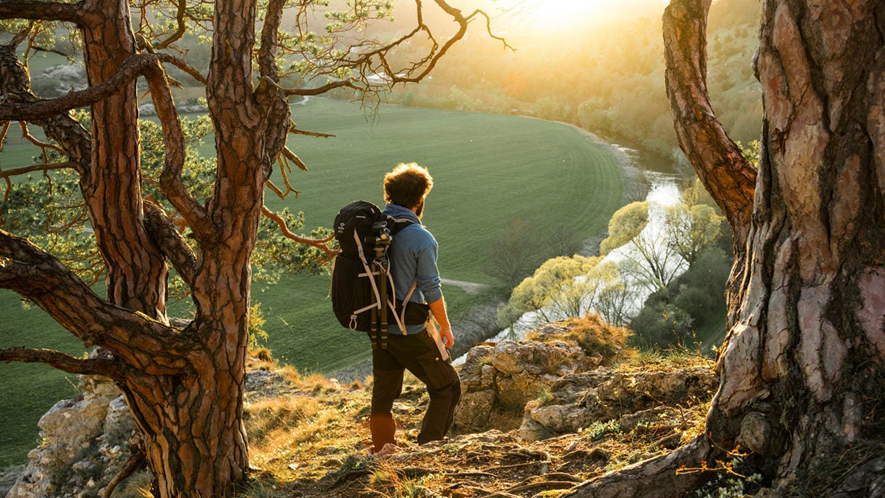Mann mit Rucksack steht an einem Hang und schaut in Richtung eines Sees